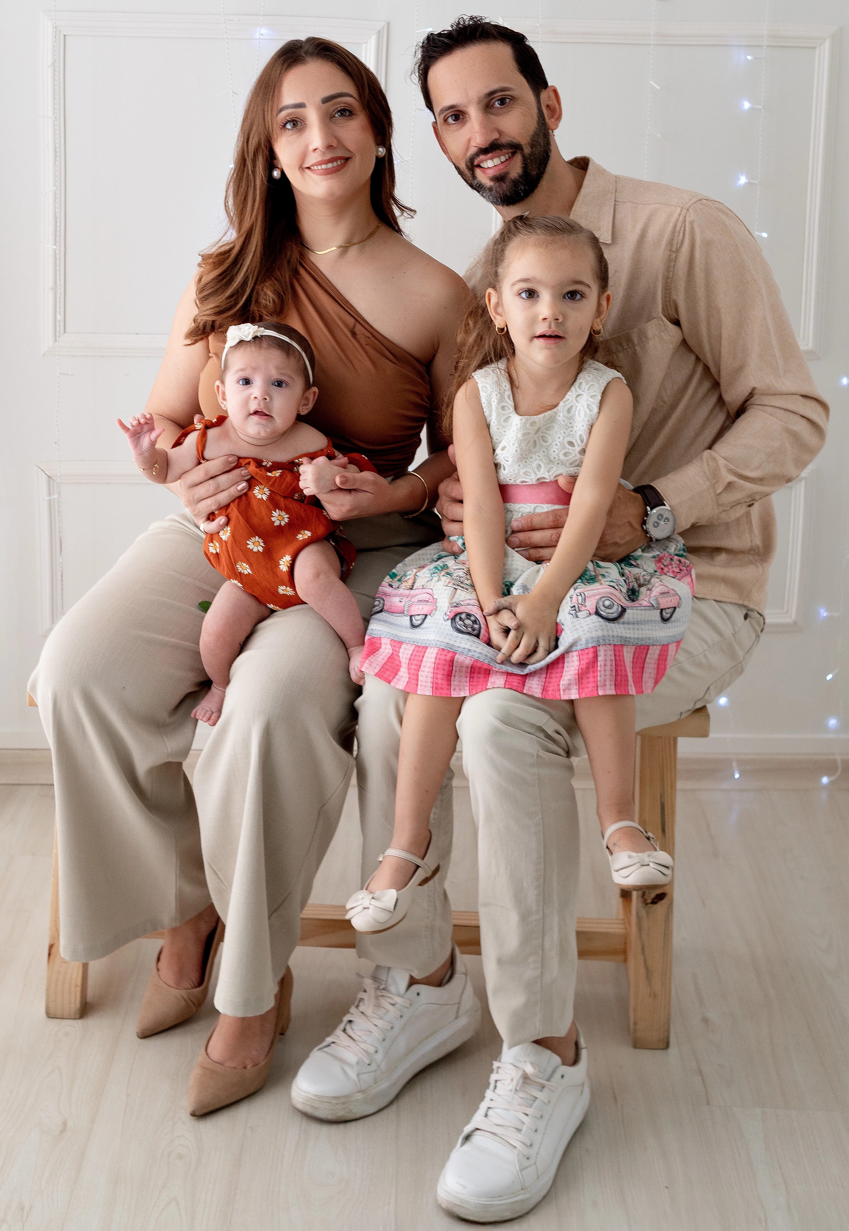 Homem e mulher sentados em um banco com suas duas filhas no colo, posando para um retrato de família em estúdio.