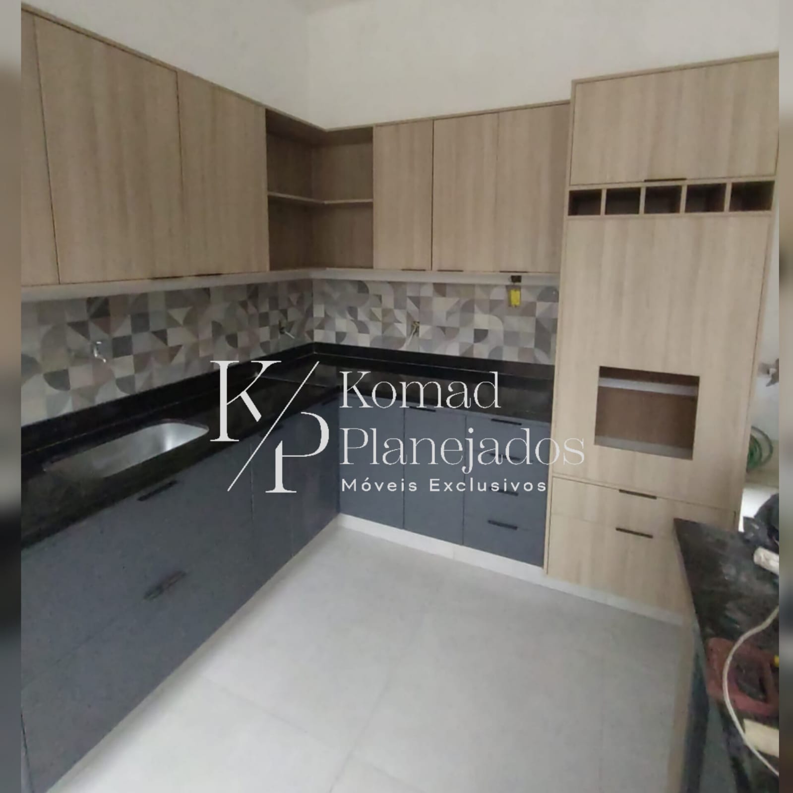Cozinha moderna com móveis planejados da Komad Planejados. Armários aéreos e torre quente em padrão madeira, gabinetes inferiores na cor cinza escuro com puxadores discretos. Bancada preta, cuba de inox e azulejos decorativos.