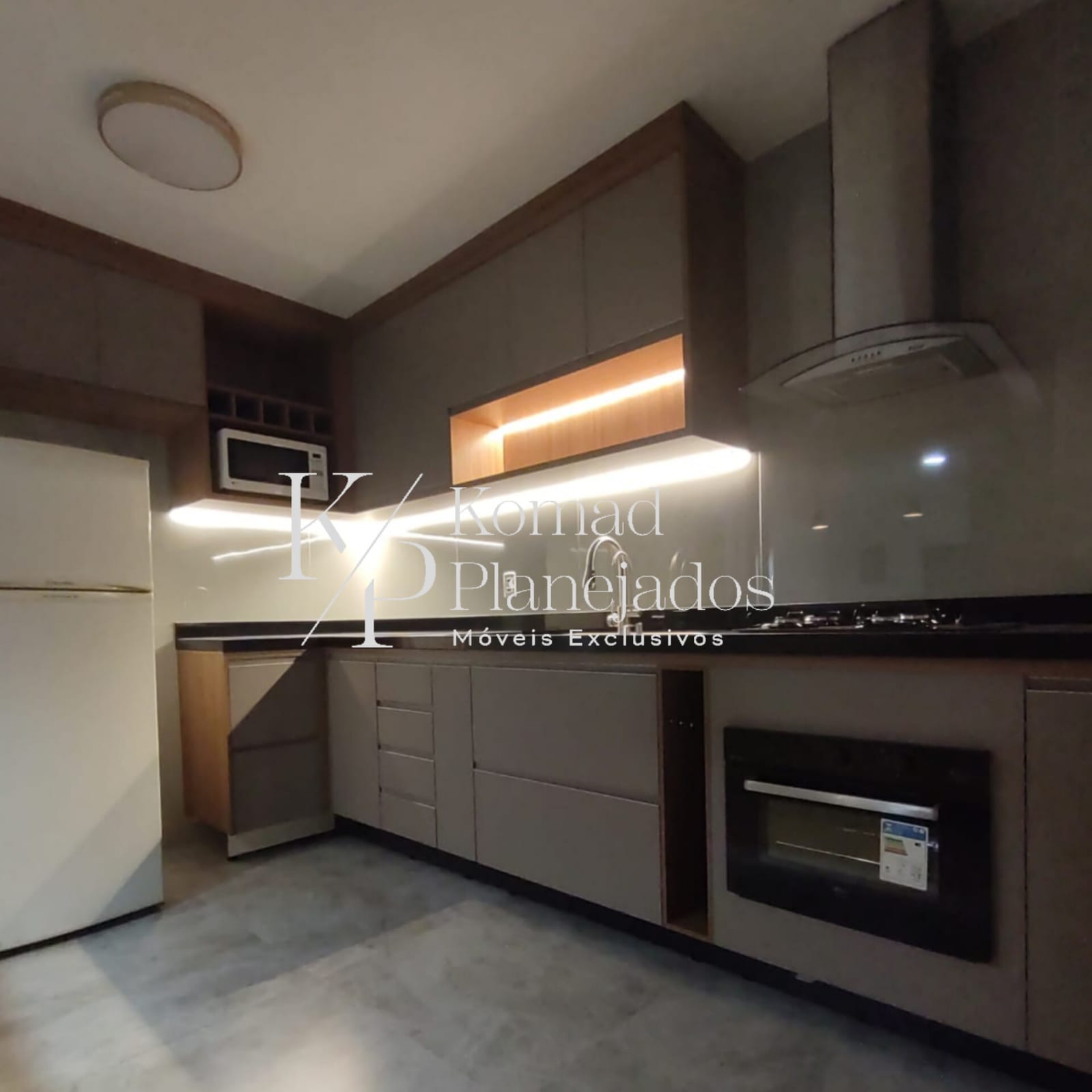 Cozinha planejada moderna com armários em tom cinza e detalhes em madeira. Bancada preta com cooktop, coifa de inox e iluminação de LED embutida sob os armários superiores, com o logo da Komad Planejados.