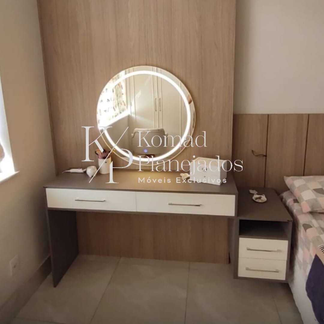 Canto de quarto com móveis planejados. Penteadeira branca e cinza com gavetas e um espelho redondo com iluminação de LED. Ao fundo, um painel de madeira que compõe a cabeceira da cama. Projeto da Komad Planejados.