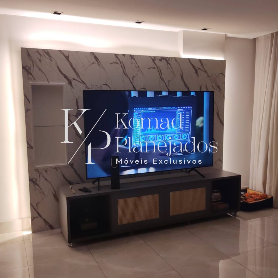 Painel de TV planejado para sala, com um grande painel ao fundo que imita mármore carrara. Rack baixo na cor grafite com detalhes em palhinha e rodízios. Projeto de móveis exclusivos da Komad Planejados.