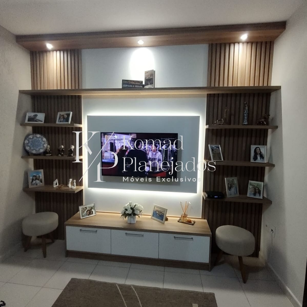 Sala de estar com painel de TV planejado da Komad Planejados. O móvel combina madeira ripada, rack branco e prateleiras de madeira para decoração. A TV está centralizada com iluminação de LED ao fundo.