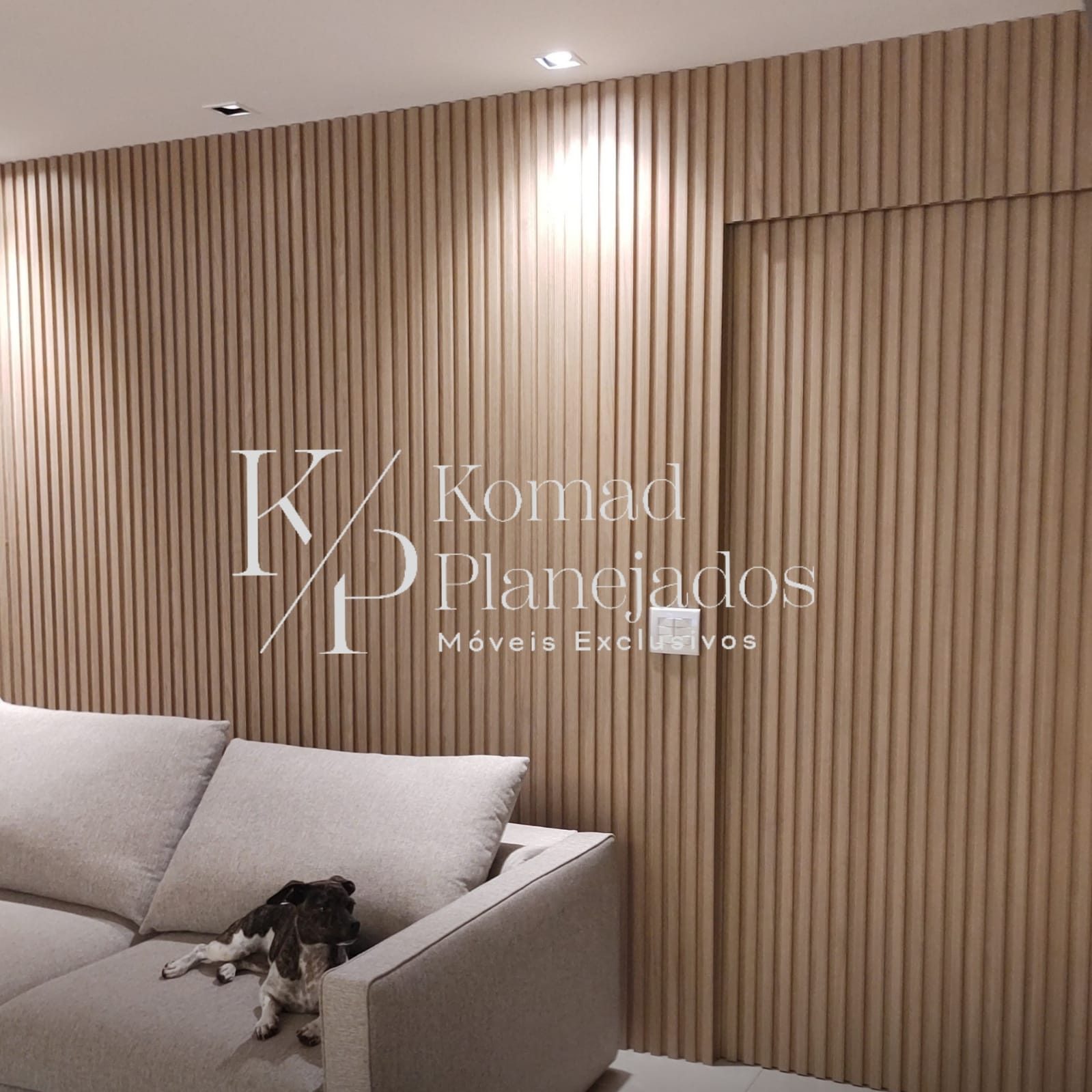 Detalhe de uma sala com um grande painel de madeira ripada que cobre toda a parede e oculta uma porta. Um sofá cinza claro e um cachorro pequeno estão em primeiro plano. Logo da Komad Planejados sobre o painel.